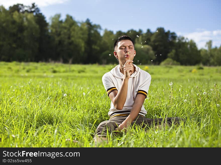 Young handsome guy on the green sunny meadow with the grass blowing on a dandelion