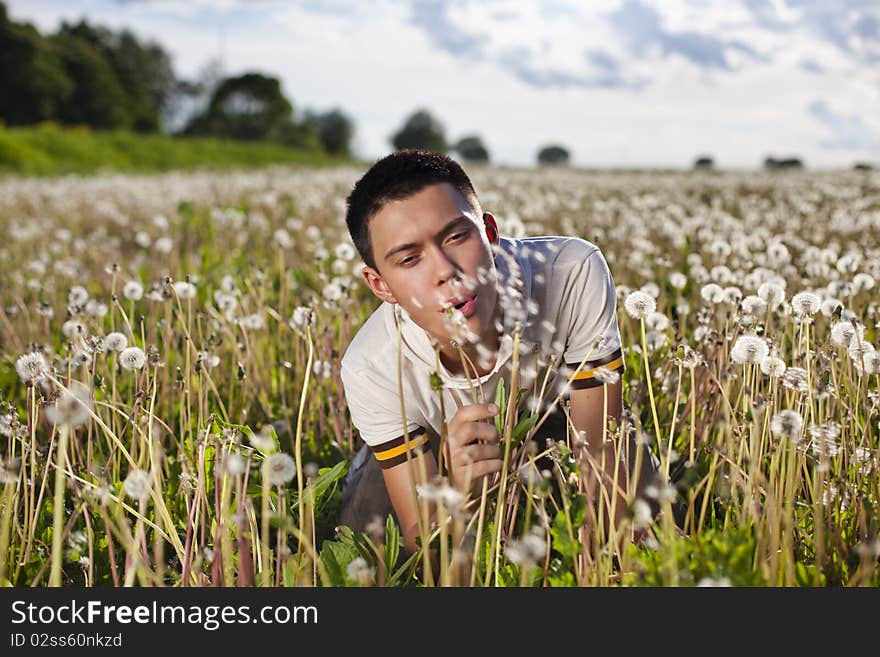 Young handsome guy on the green sunny meadow with the grass blowing on a dandelion