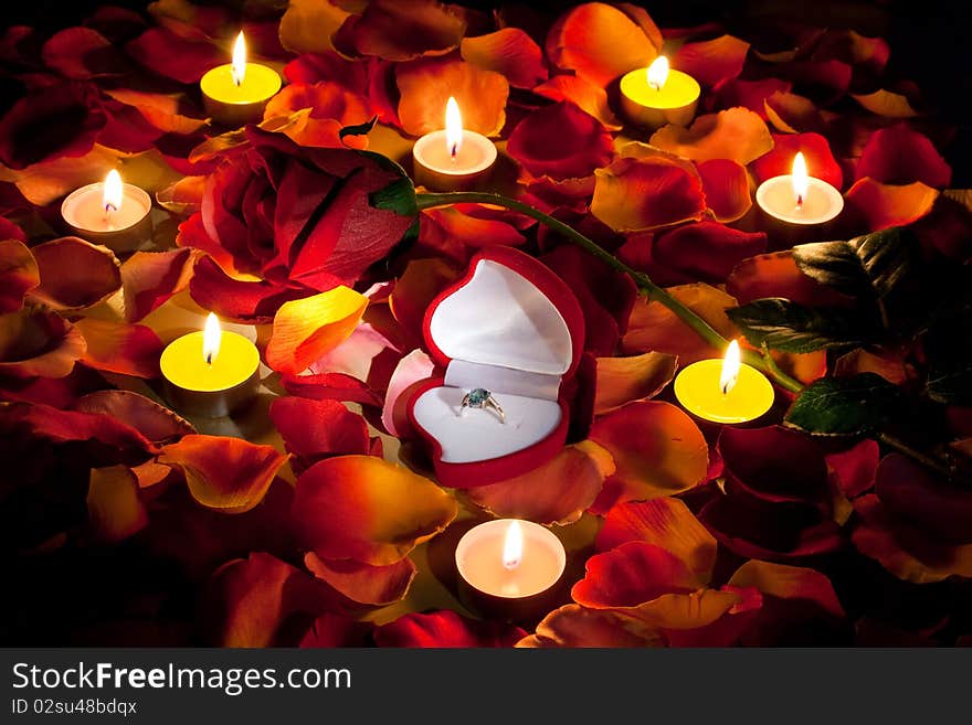 The professions of love represented by the rose petals with ring in box and red rose lit up by candles on the table.