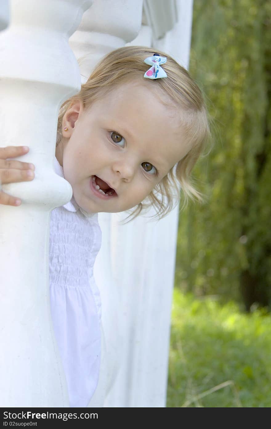 The girl in a white dress with a hairpin in hair looks out because of a column and rejoices. The girl in a white dress with a hairpin in hair looks out because of a column and rejoices