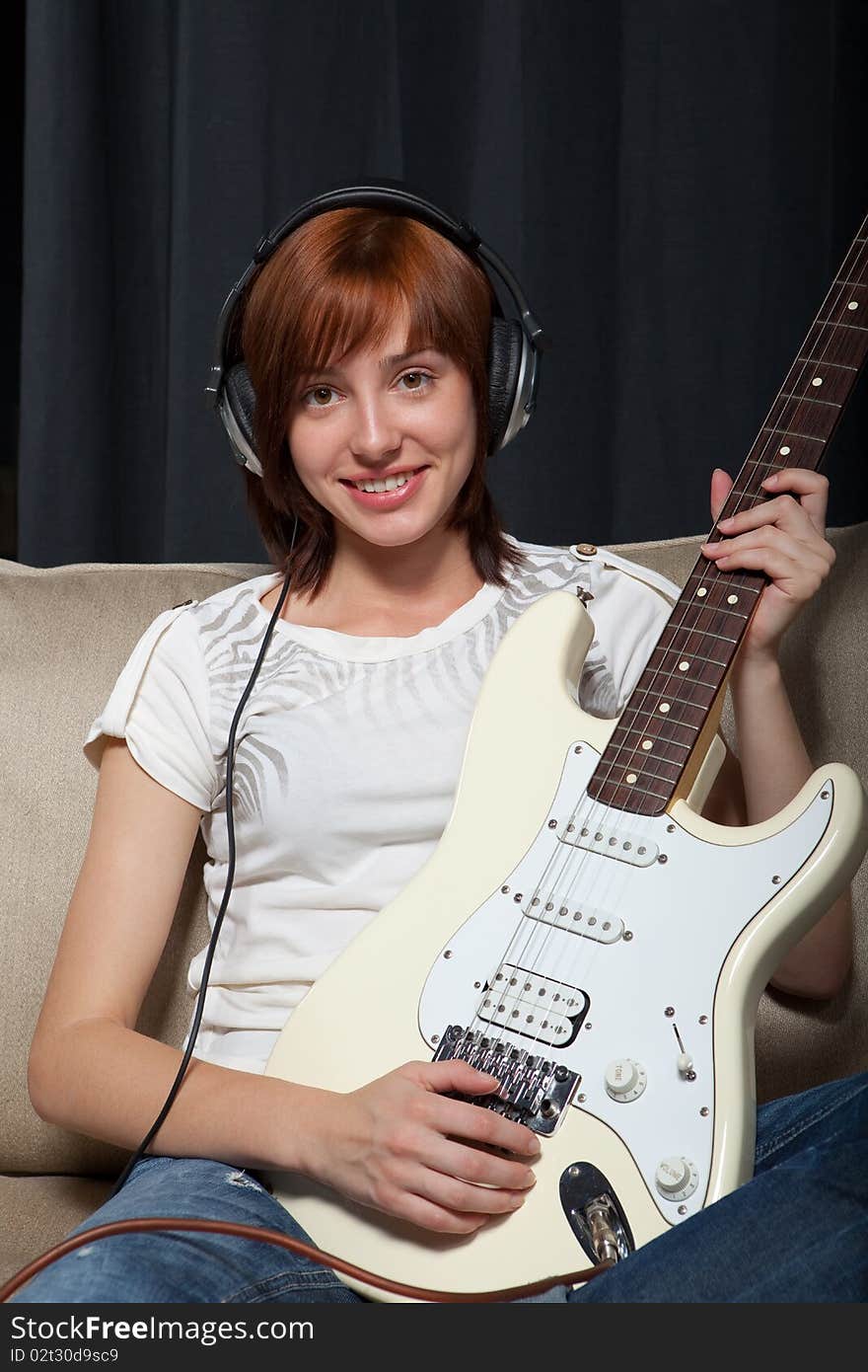 Teenage girl sitting on the sofa in headphones playing electric guitar.