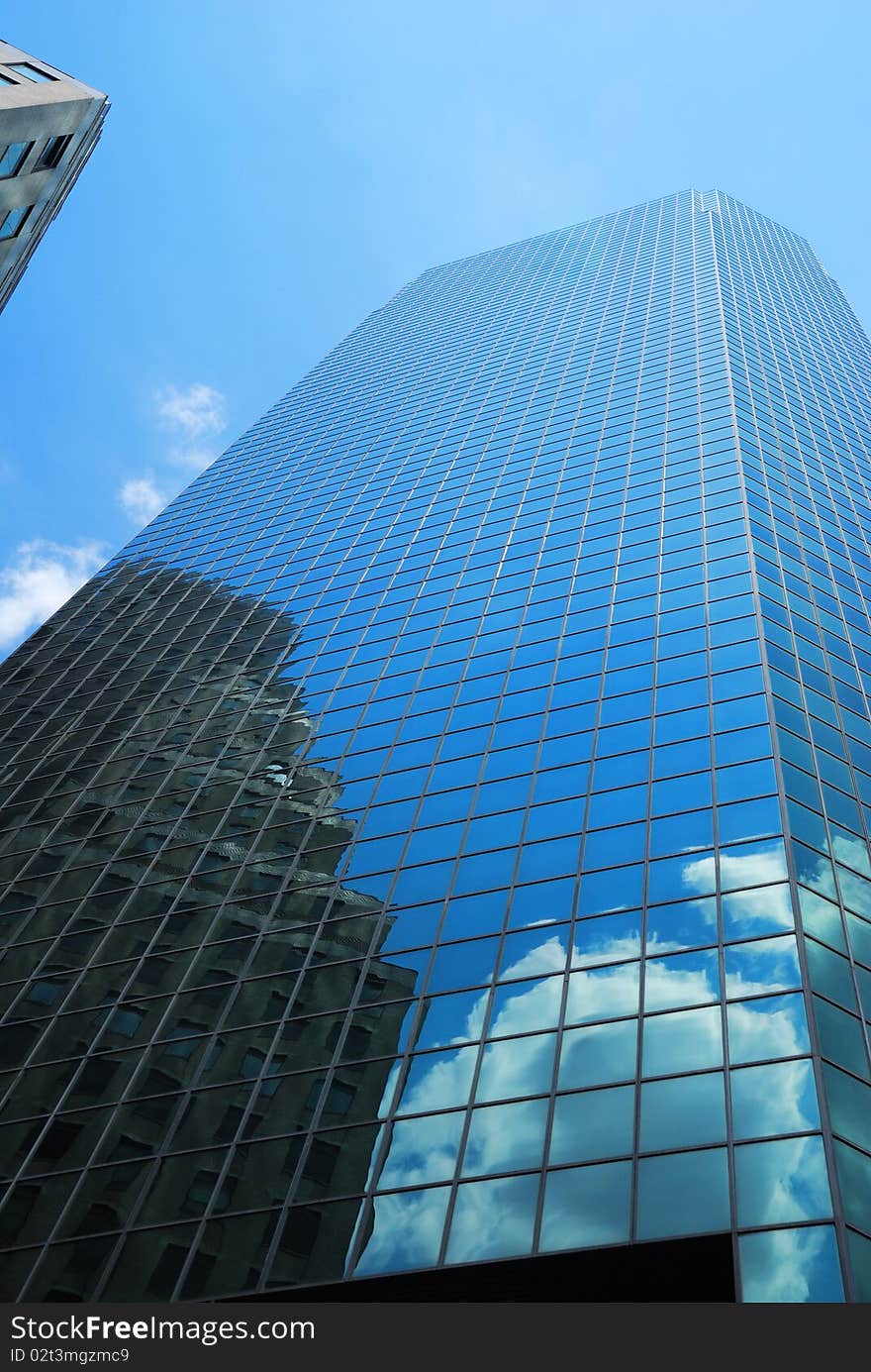 Glassy skyscraper of New York is photographed from below against blue sky. Next building and white cloud are reflected in its mirror surface. Glassy skyscraper of New York is photographed from below against blue sky. Next building and white cloud are reflected in its mirror surface.