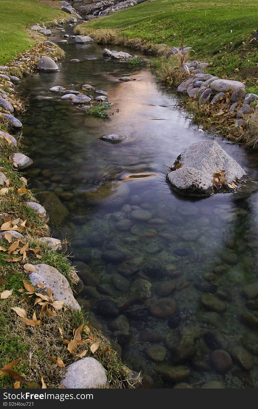 A slow moving creek with green grass and fall leaves glows as the sun rises. A slow moving creek with green grass and fall leaves glows as the sun rises.
