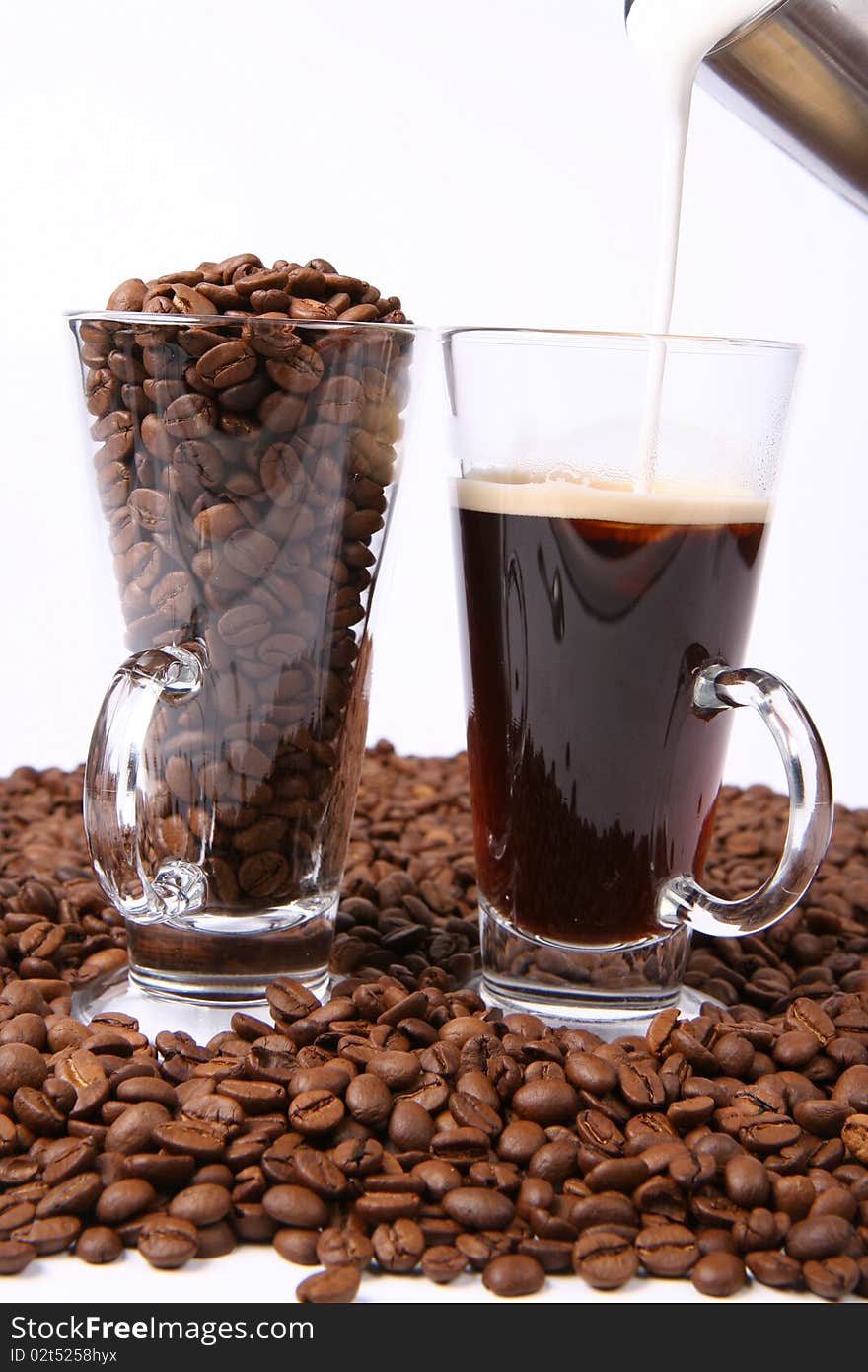 Making of caffe latte:glass of black coffee with milk being poured into it, with a glass of beans surrounded by coffee beans