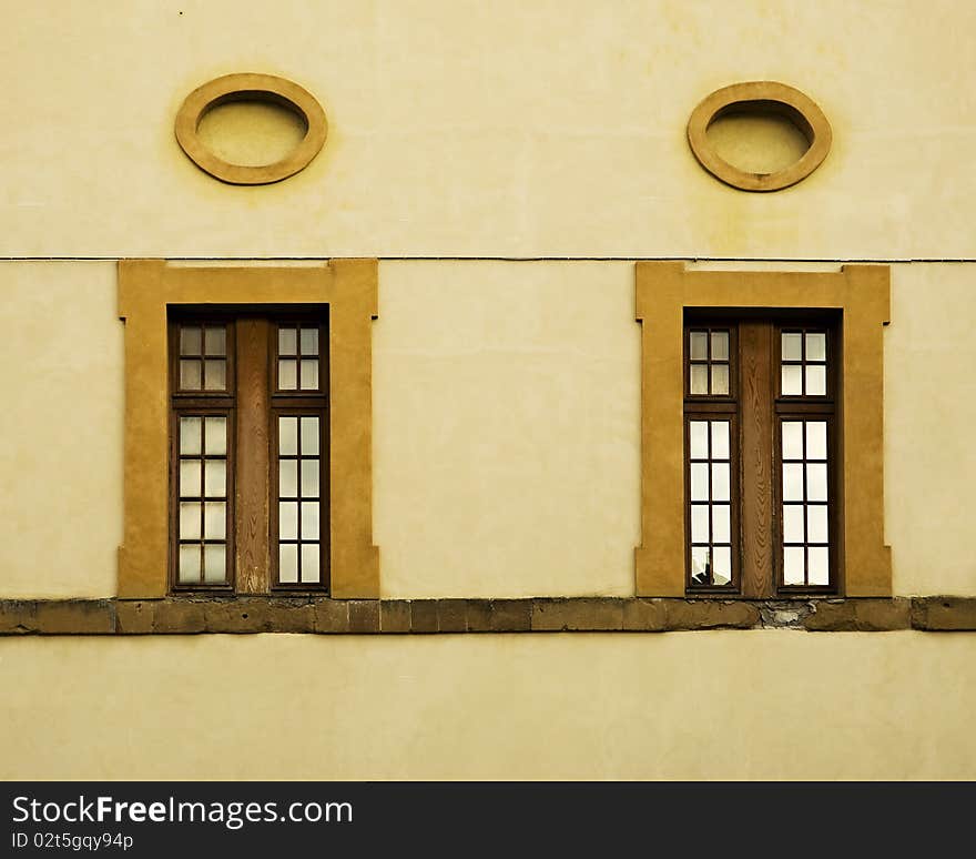 A yellow wall in Italy, with brown windows, deep yellow window frames and ovals above the windows, and a line of stonework. A yellow wall in Italy, with brown windows, deep yellow window frames and ovals above the windows, and a line of stonework.