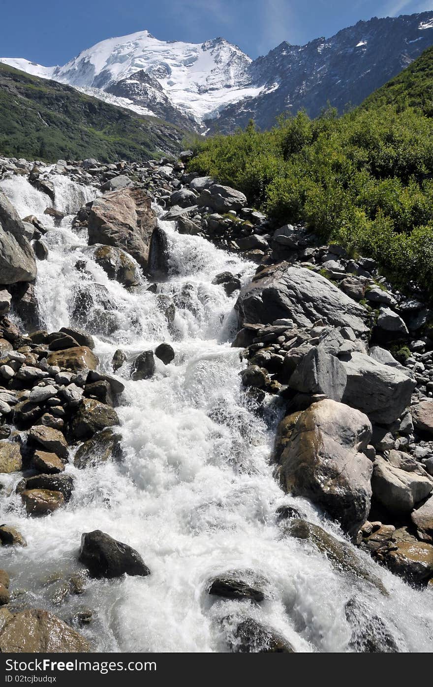 View of rapid stream and high mountains with snow in background.