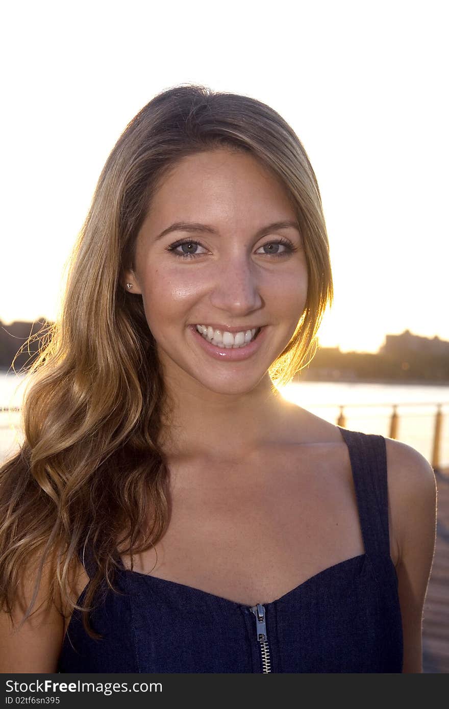 A young blond woman, dressed in a blue sun dress, smiles brightly on a pier at dusk. A young blond woman, dressed in a blue sun dress, smiles brightly on a pier at dusk