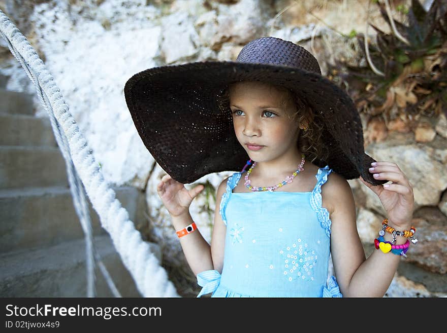 Outdoor portrait of a beautiful little girl with curly hairs in a wide hat on rock background. shot made on a shore of the sea. Outdoor portrait of a beautiful little girl with curly hairs in a wide hat on rock background. shot made on a shore of the sea