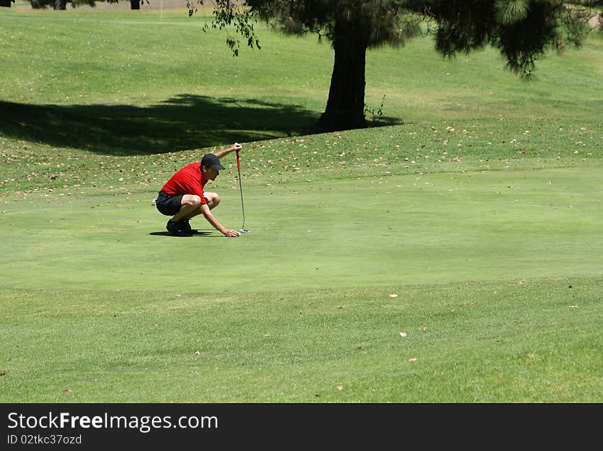 Man wearing a red shirt back cap and shorts on the green lining up his shot to get the golf ball in the hole. Man wearing a red shirt back cap and shorts on the green lining up his shot to get the golf ball in the hole