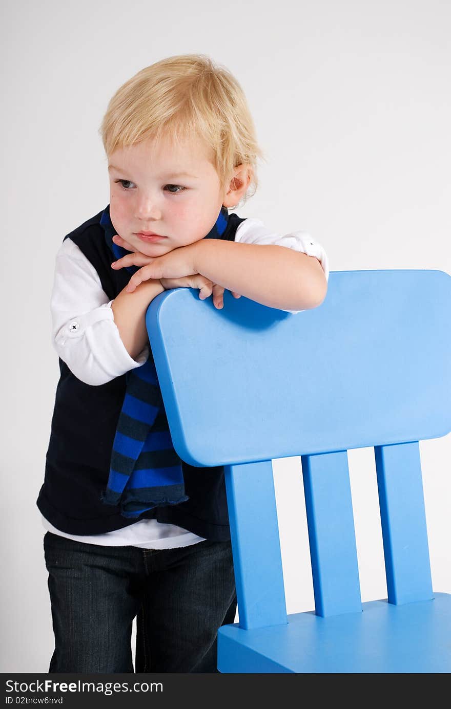 A vertical image of a handsome young blonde boy leaning on a blue chair deep in thought. A vertical image of a handsome young blonde boy leaning on a blue chair deep in thought