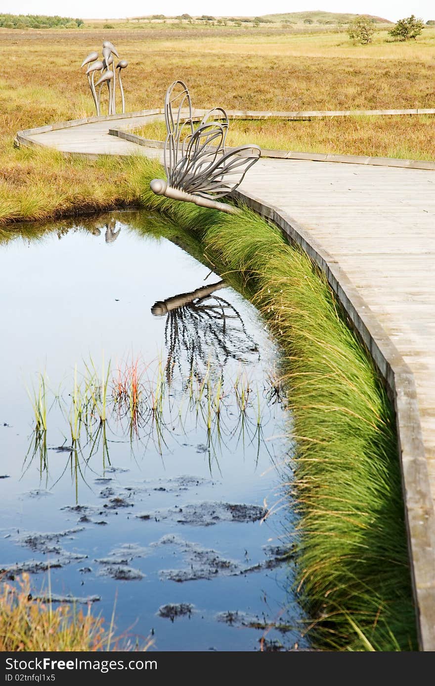 A vertical image of a wooden boardwalk beside a pond with a dragonfly ornament. A vertical image of a wooden boardwalk beside a pond with a dragonfly ornament