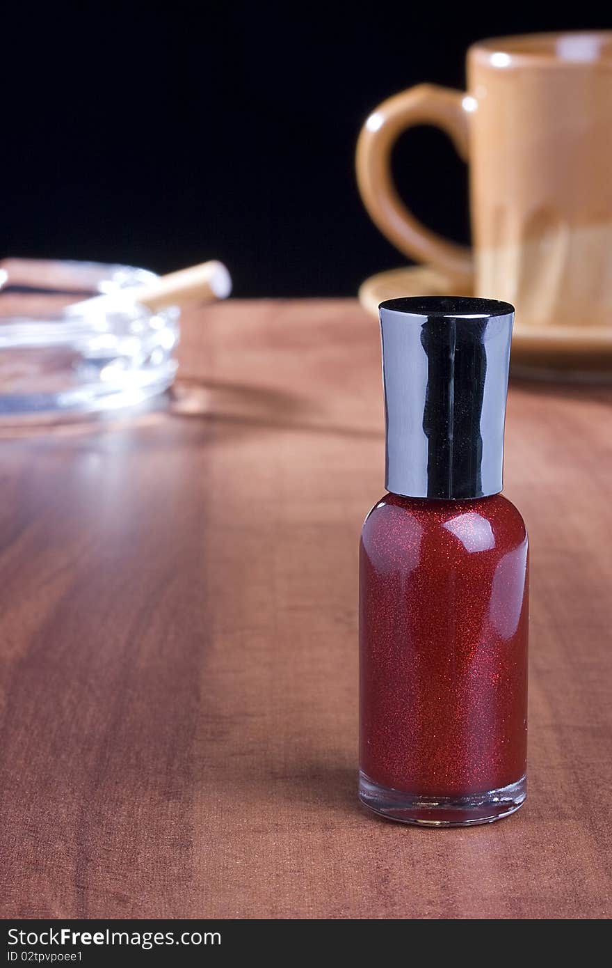 Red nail polish standing on a coffee table in front of a cup of coffee and an ashtray.