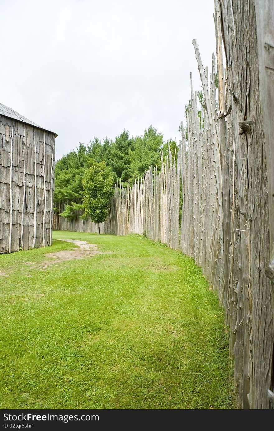 Walkway between a high fence/wall made of bare tree trunks and a long house. Walkway between a high fence/wall made of bare tree trunks and a long house.