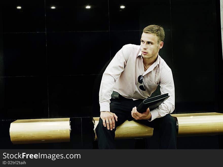 The attractive young man with a folder sits on a bench and expects a meeting. The attractive young man with a folder sits on a bench and expects a meeting.