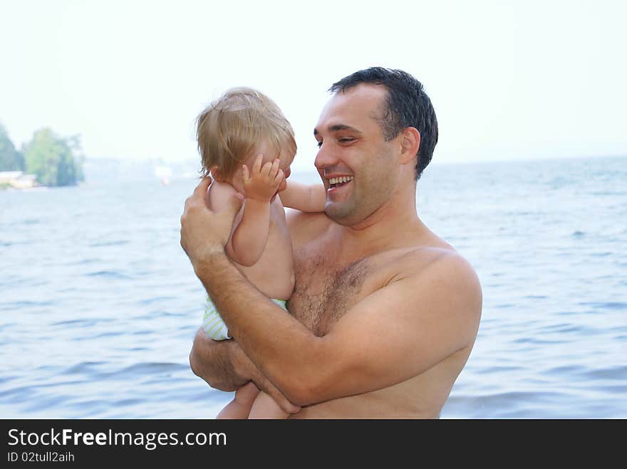 In the summer on a beach the laughing father holds on hands of the small child and plays with it. In the summer on a beach the laughing father holds on hands of the small child and plays with it