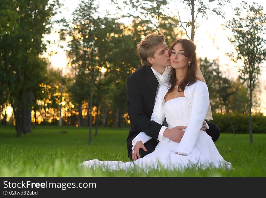 The groom kisses the bride sitting on a lawn in park