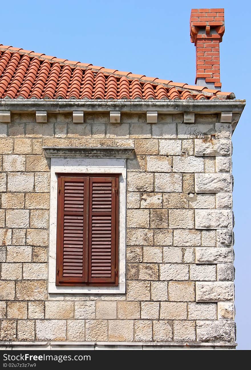 Part of the old stone building. Closeup of the window with shutters on the facade, red tiled roof and bricked chimney. Part of the old stone building. Closeup of the window with shutters on the facade, red tiled roof and bricked chimney.