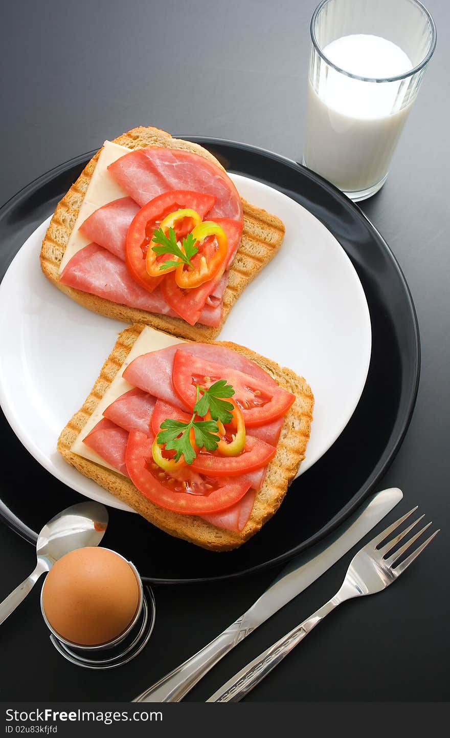 Pork tenderloin sandwiches, cheese, tomato and pepper arranged in ceramic plates in the company of soft-boiled egg placed in a special metal stand, fork, knife, spoon and glass of milk. Pork tenderloin sandwiches, cheese, tomato and pepper arranged in ceramic plates in the company of soft-boiled egg placed in a special metal stand, fork, knife, spoon and glass of milk