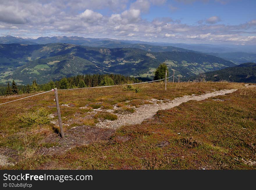 Panoramic view from mountain Gerlitzen to Carinthia, Austria. Image shows a hiking trail with meadow and a fence. In the background is the landscape of Carinthia, Austria. Sky is blue and cloudy. Panoramic view from mountain Gerlitzen to Carinthia, Austria. Image shows a hiking trail with meadow and a fence. In the background is the landscape of Carinthia, Austria. Sky is blue and cloudy.