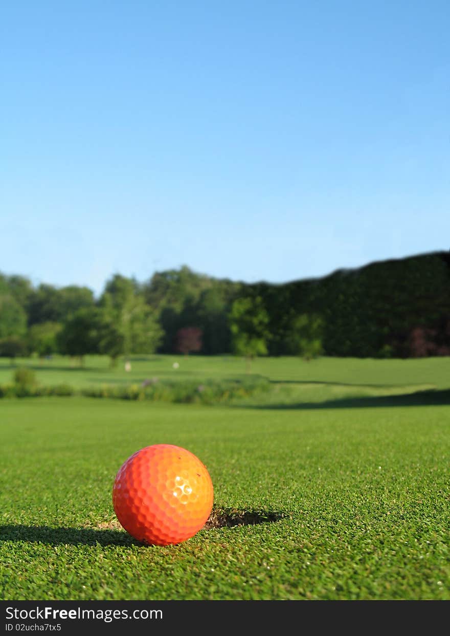 Orange gold ball on the green beside the hole with clear blue sky in background.