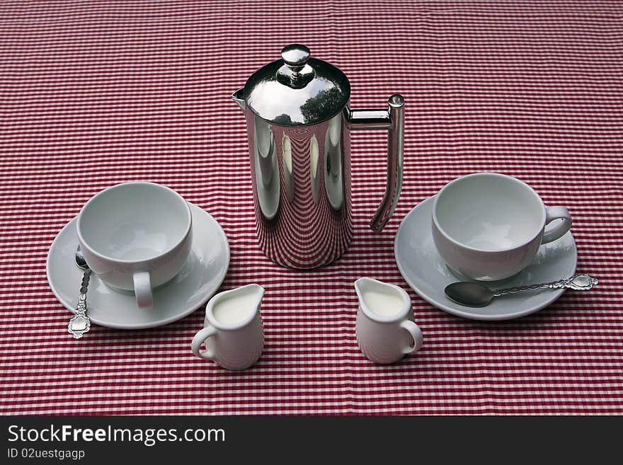 A shiny coffee pot and cups and saucers on a red and white tablecloth