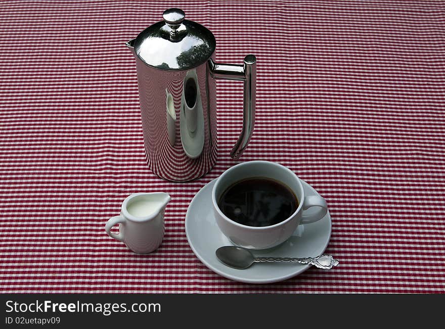 A shiny coffee pot and cups and saucers on a red and white tablecloth