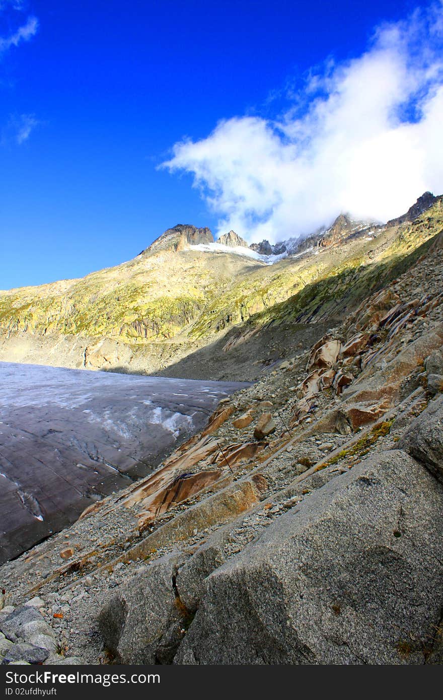 A rocky mountain side with snow cappe mountains and a glacier with clouds and a bright blue sunny sky. A rocky mountain side with snow cappe mountains and a glacier with clouds and a bright blue sunny sky