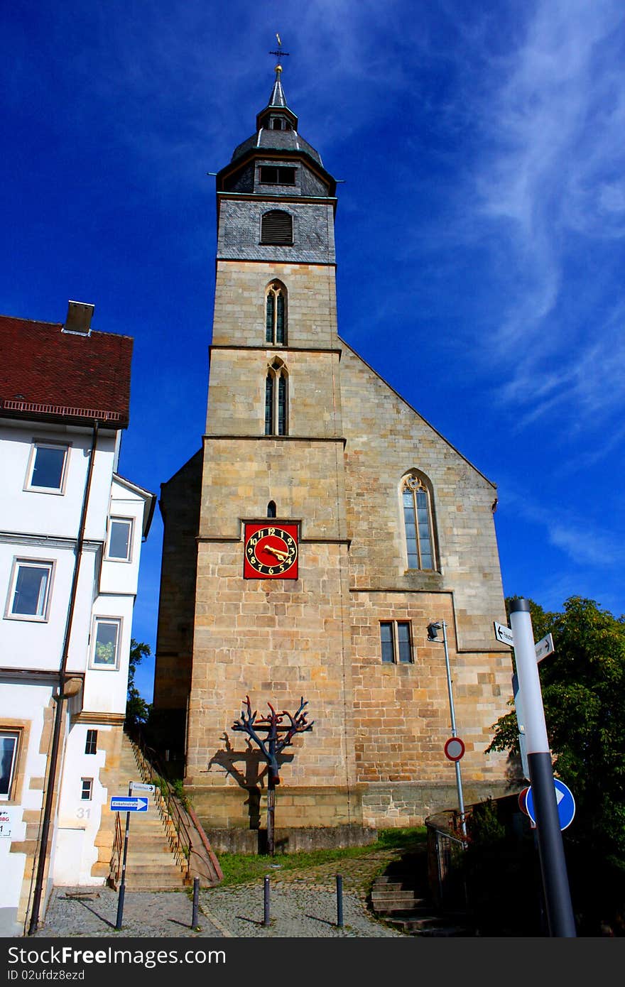 A old church in with a large steeple under a blue sky with a wooden cross and a red clock in Stuttgart in Germany. A old church in with a large steeple under a blue sky with a wooden cross and a red clock in Stuttgart in Germany