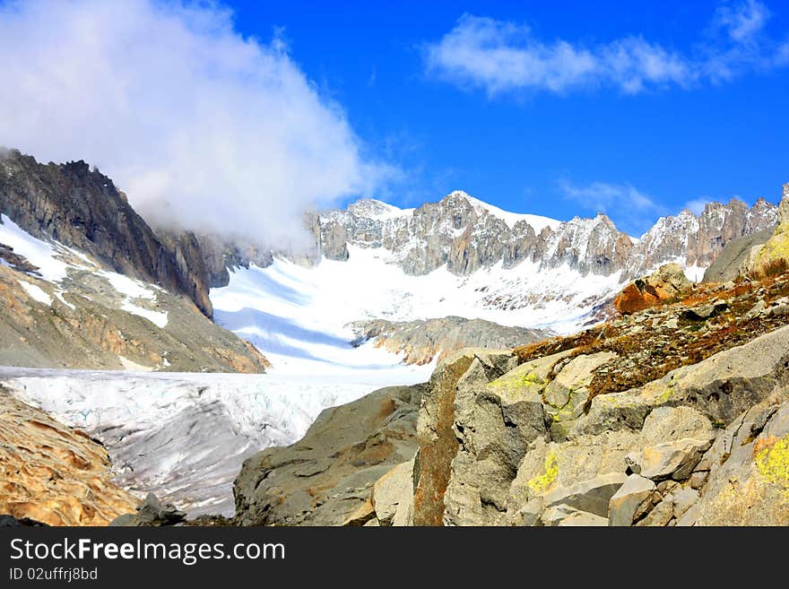 Rocky mountain side covering a glacier with snowy mountains in the back under a blue sunny sky and clouds creeping in. Rocky mountain side covering a glacier with snowy mountains in the back under a blue sunny sky and clouds creeping in