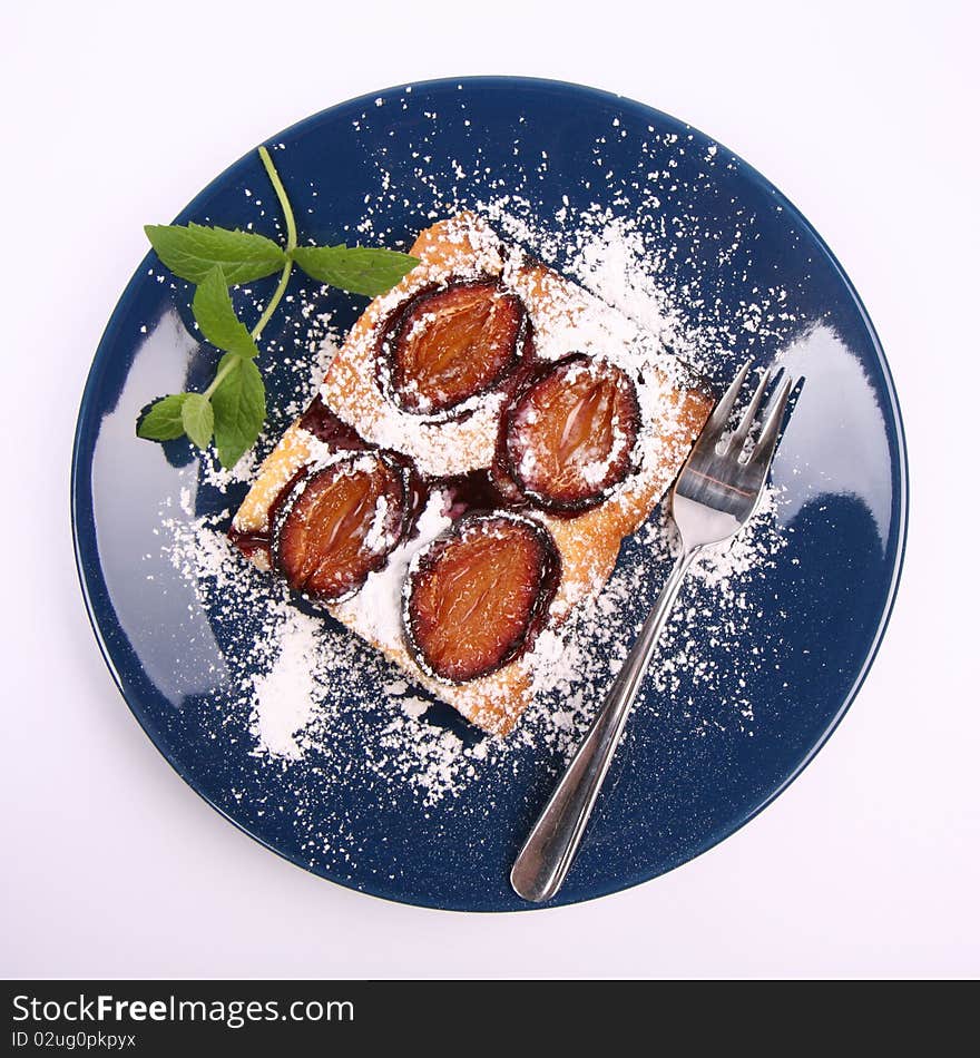 Piece of Plum Pie covered with powder sugar on a blue plate decorated with a mint twig