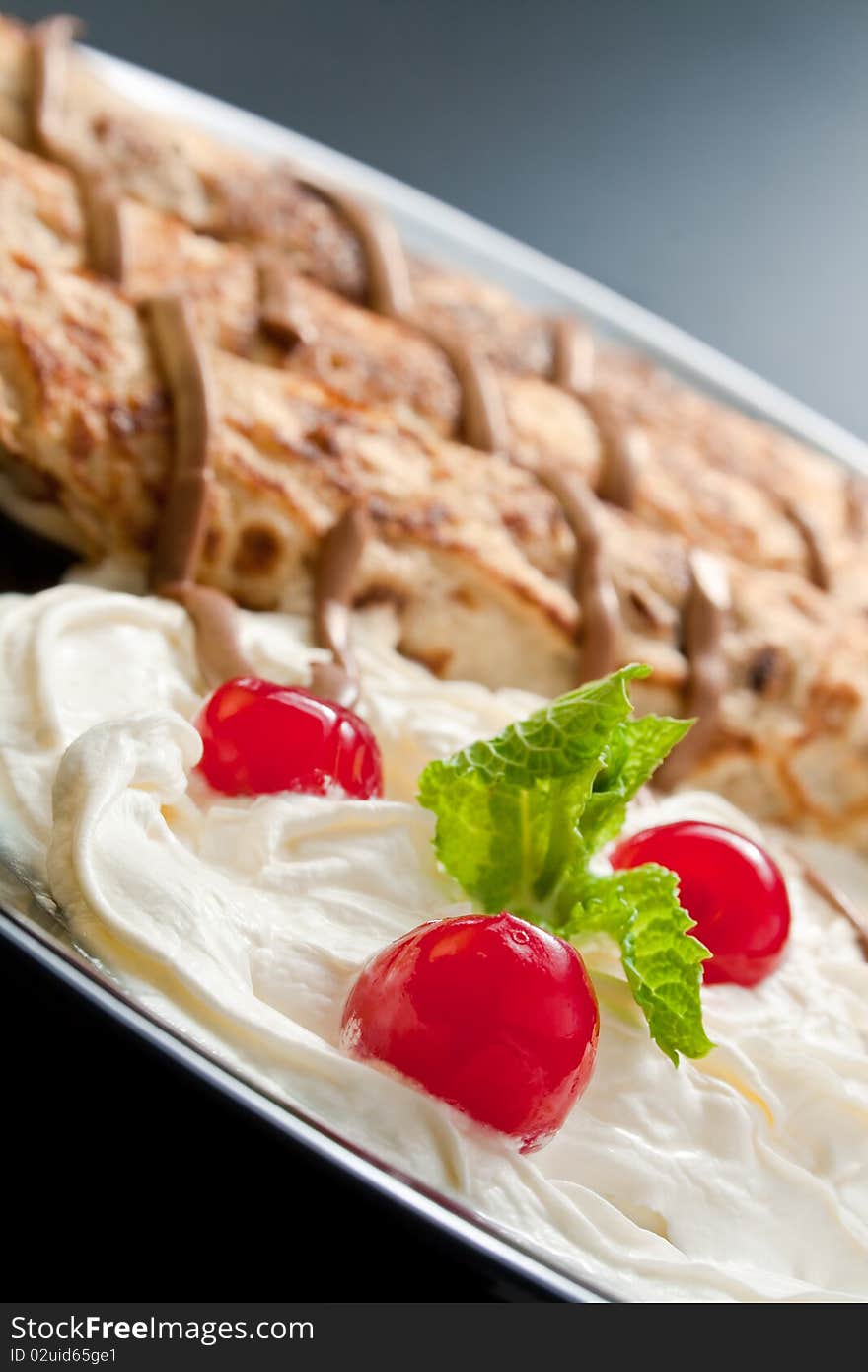 Three rolled pancakes close-up arranged with cream, cherries, chocolate and sprig mint leaves in black ceramic plate. Three rolled pancakes close-up arranged with cream, cherries, chocolate and sprig mint leaves in black ceramic plate