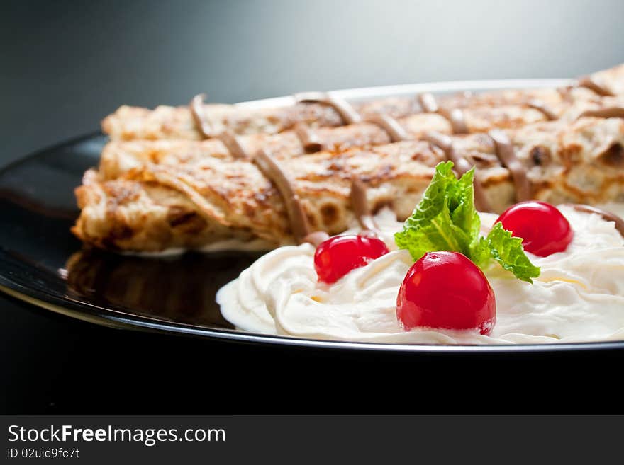 Three rolled pancakes close-up arranged with cream, cherries, chocolate and sprig mint leaves in black ceramic plate. Three rolled pancakes close-up arranged with cream, cherries, chocolate and sprig mint leaves in black ceramic plate