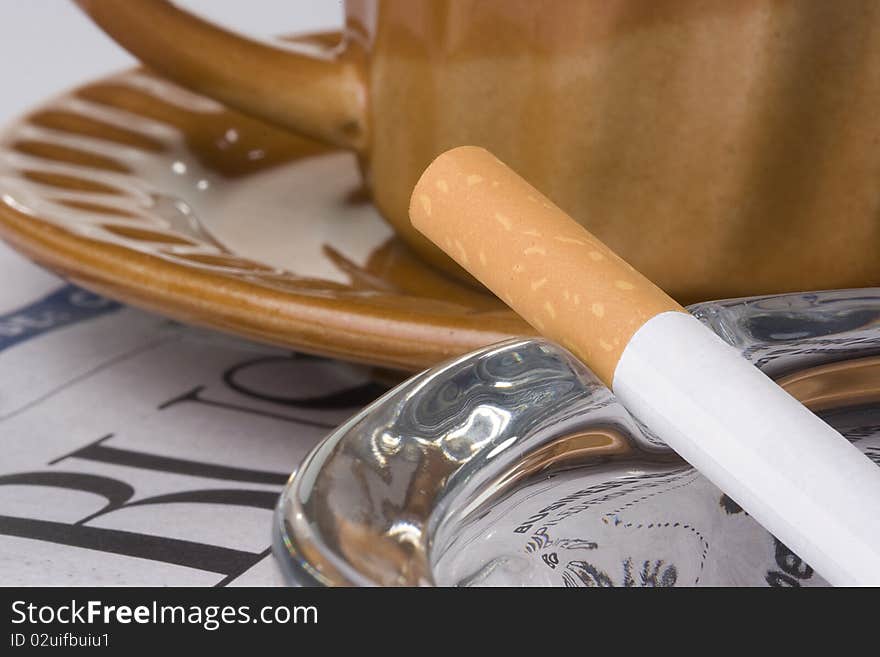 A filtered cigarette resting on a glass ashtray next to a brown cup and plate and a newspaper. A filtered cigarette resting on a glass ashtray next to a brown cup and plate and a newspaper.