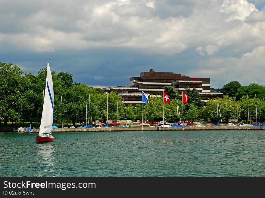 Sailboat coming from Zurich yachtport, deep turquoise water of the zurich lake and some yachts are at foreground and the sky conered with rainy clouds is at background