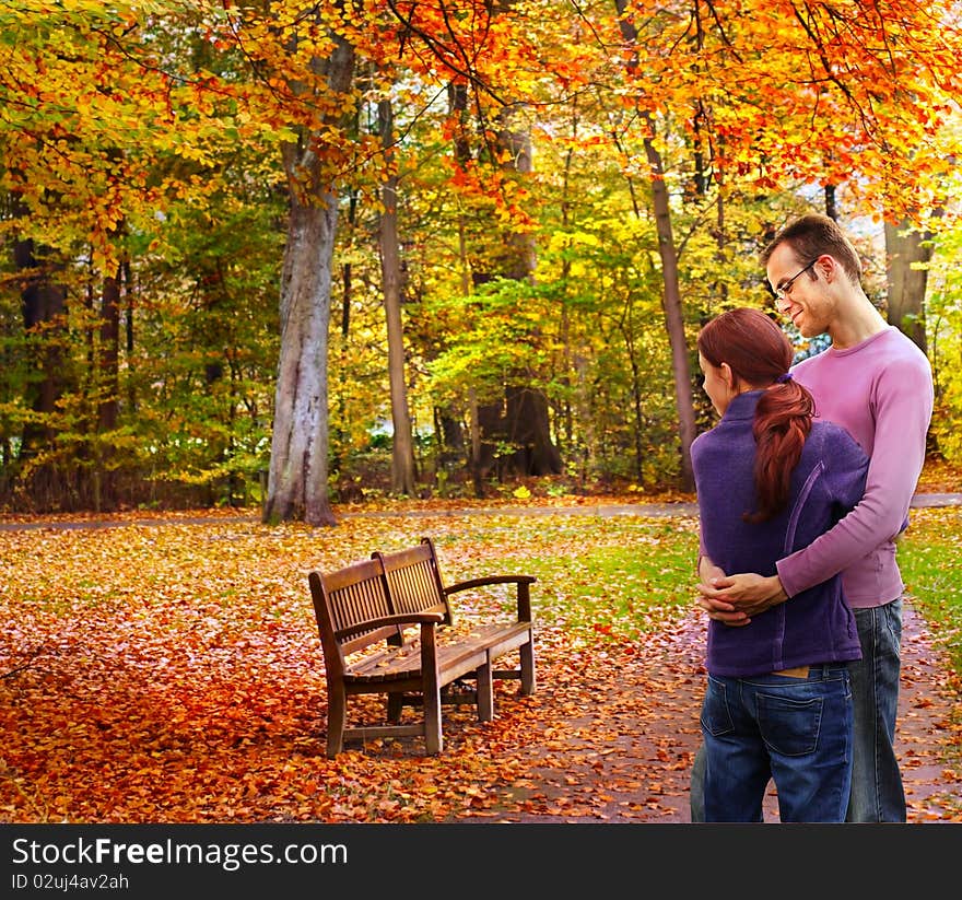 Happy couple standing embraced in the autumnal park looking to a bench. Happy couple standing embraced in the autumnal park looking to a bench