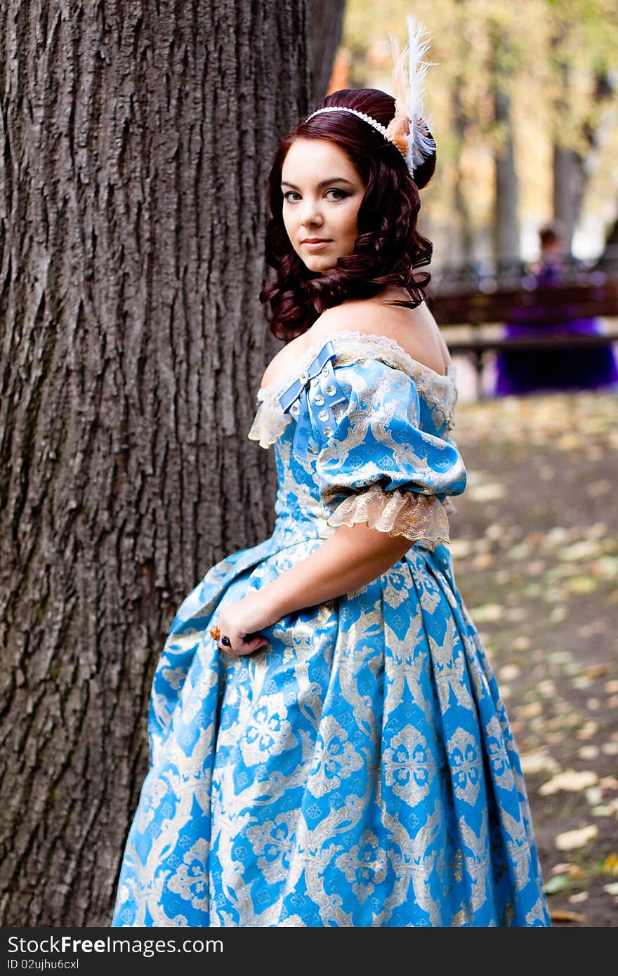 A portrait of lady in a blue baroque dress standing near a bole. A portrait of lady in a blue baroque dress standing near a bole