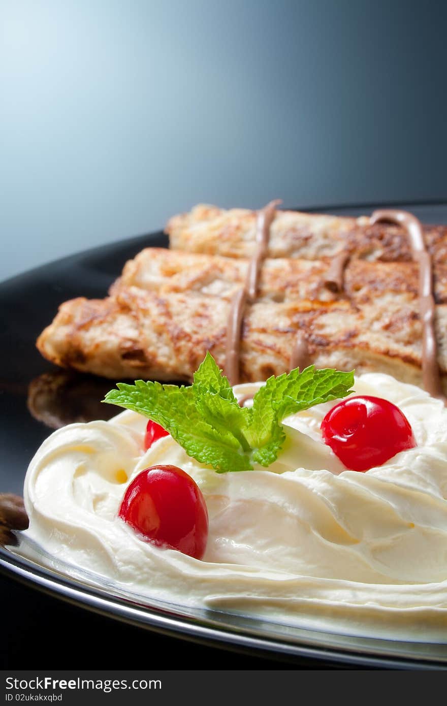 Three rolled pancakes close-up arranged with cream, cherries, chocolate and sprig mint leaves in black ceramic plate. Three rolled pancakes close-up arranged with cream, cherries, chocolate and sprig mint leaves in black ceramic plate