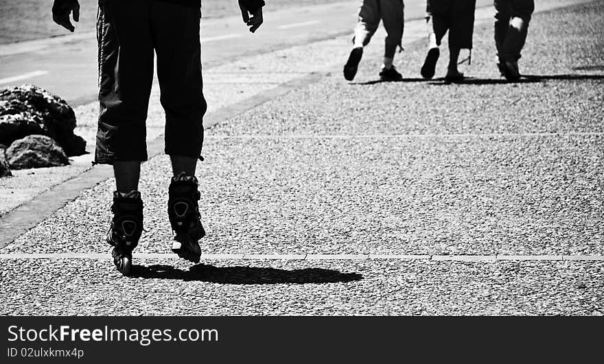 A little boy is sketting in the street we can see just his legs an a part of his hands. three persons are walking in front of his. The image is in black and white. A little boy is sketting in the street we can see just his legs an a part of his hands. three persons are walking in front of his. The image is in black and white.