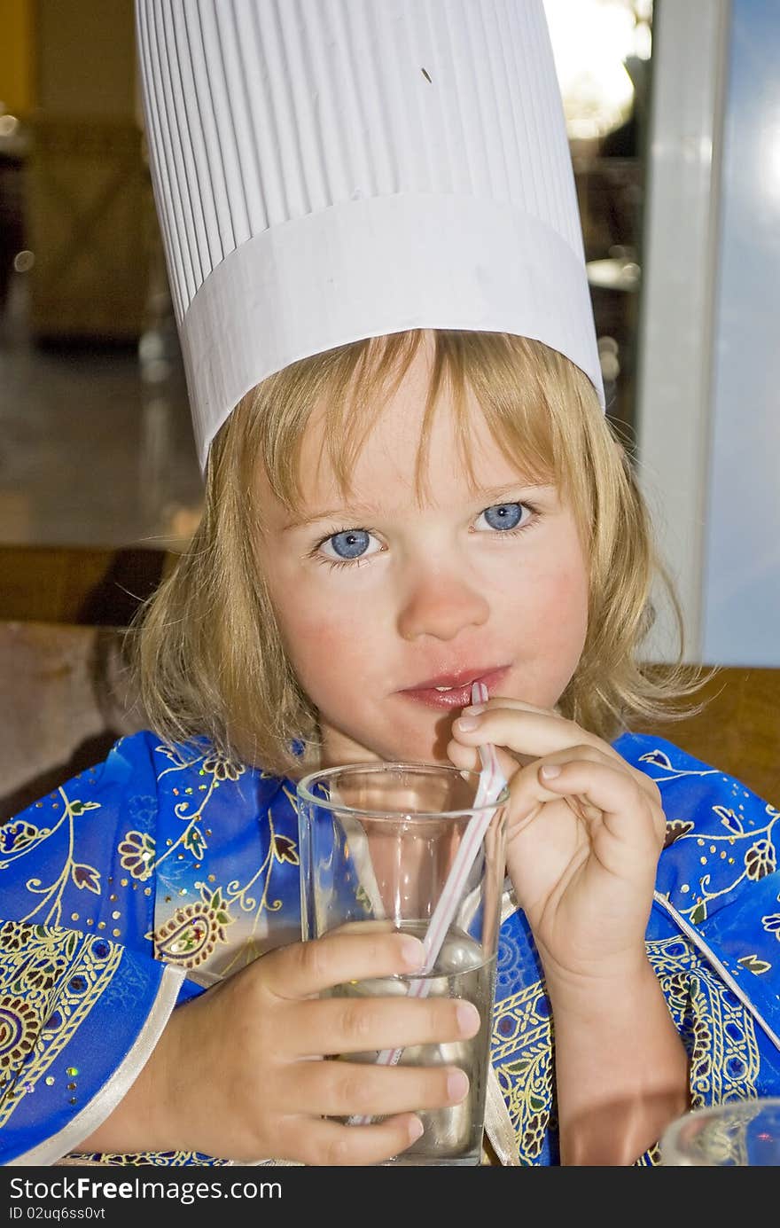 Little beautiful girl in blue dress and cook hat drinking water in stow. Little beautiful girl in blue dress and cook hat drinking water in stow.