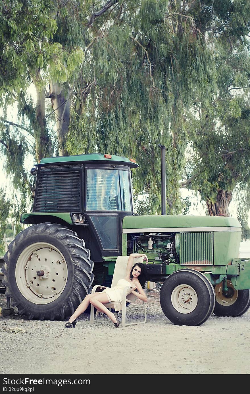 Country farm girl, beautiful young woman wearing a dress sitting on chair beside tractor