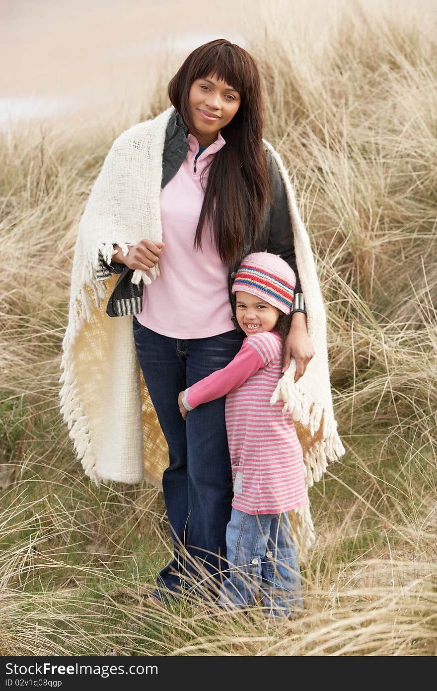 Mother And Daughter Wrapped In Blanket Amongst Sand Dunes On Winter Beach Smiling. Mother And Daughter Wrapped In Blanket Amongst Sand Dunes On Winter Beach Smiling