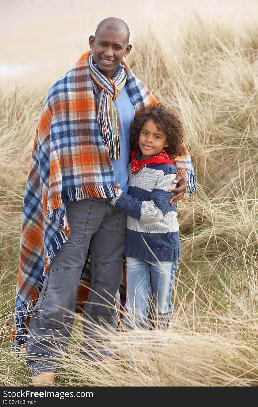 Father And Son Wrapped In Blanket Amongst Sand Dunes On Winter Beach Smiling. Father And Son Wrapped In Blanket Amongst Sand Dunes On Winter Beach Smiling