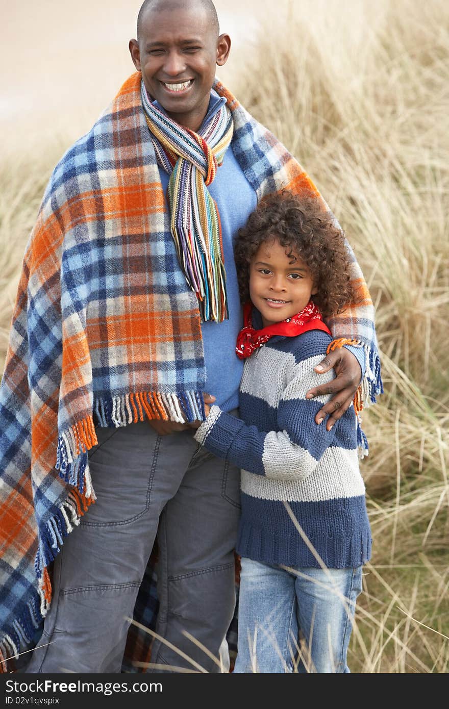 Father And Son Wrapped In Blanket Amongst Sand Dunes On Winter Beach Smiling. Father And Son Wrapped In Blanket Amongst Sand Dunes On Winter Beach Smiling