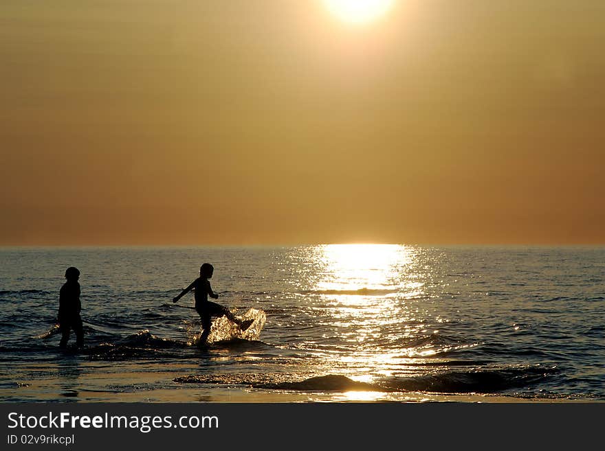Two silhouetted kids kick,play and splash in the sparkling reflection on the blue rippled water, hazy orange sky by half sunlight, warm colors, soft waves, heat from sun and cool splashing of water.