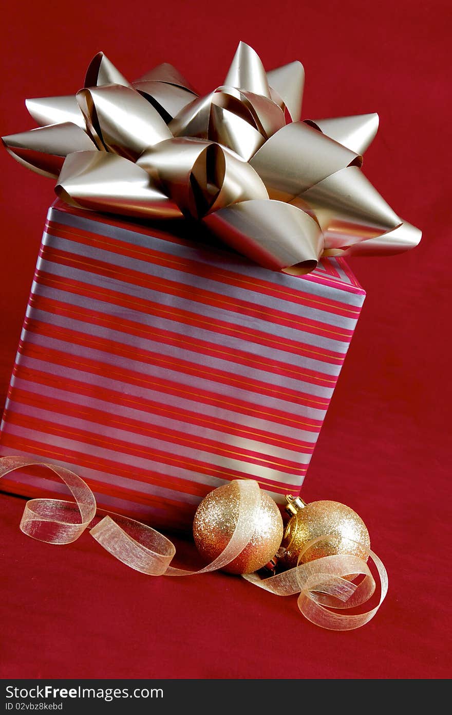 A package wrapped in silver and red paper is adorned with a large silver bow. Gold frosted ornaments and transparant gold ribbon rest in front of the package. All objects are sitting atop a red velvet background. A package wrapped in silver and red paper is adorned with a large silver bow. Gold frosted ornaments and transparant gold ribbon rest in front of the package. All objects are sitting atop a red velvet background.