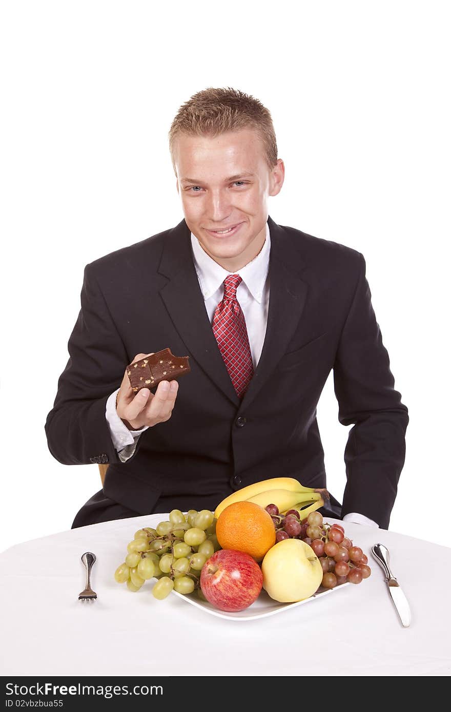 A man deciding to eat a brownie instead of his fruit on a plate in front of him. A man deciding to eat a brownie instead of his fruit on a plate in front of him.