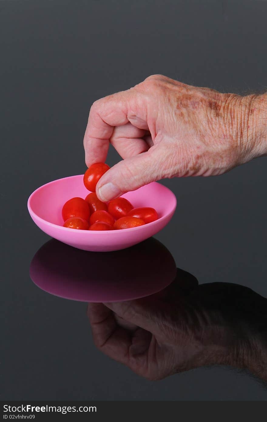 Mature male hand picking up cherry tomato from pink bowl on a black plexiglas  table.