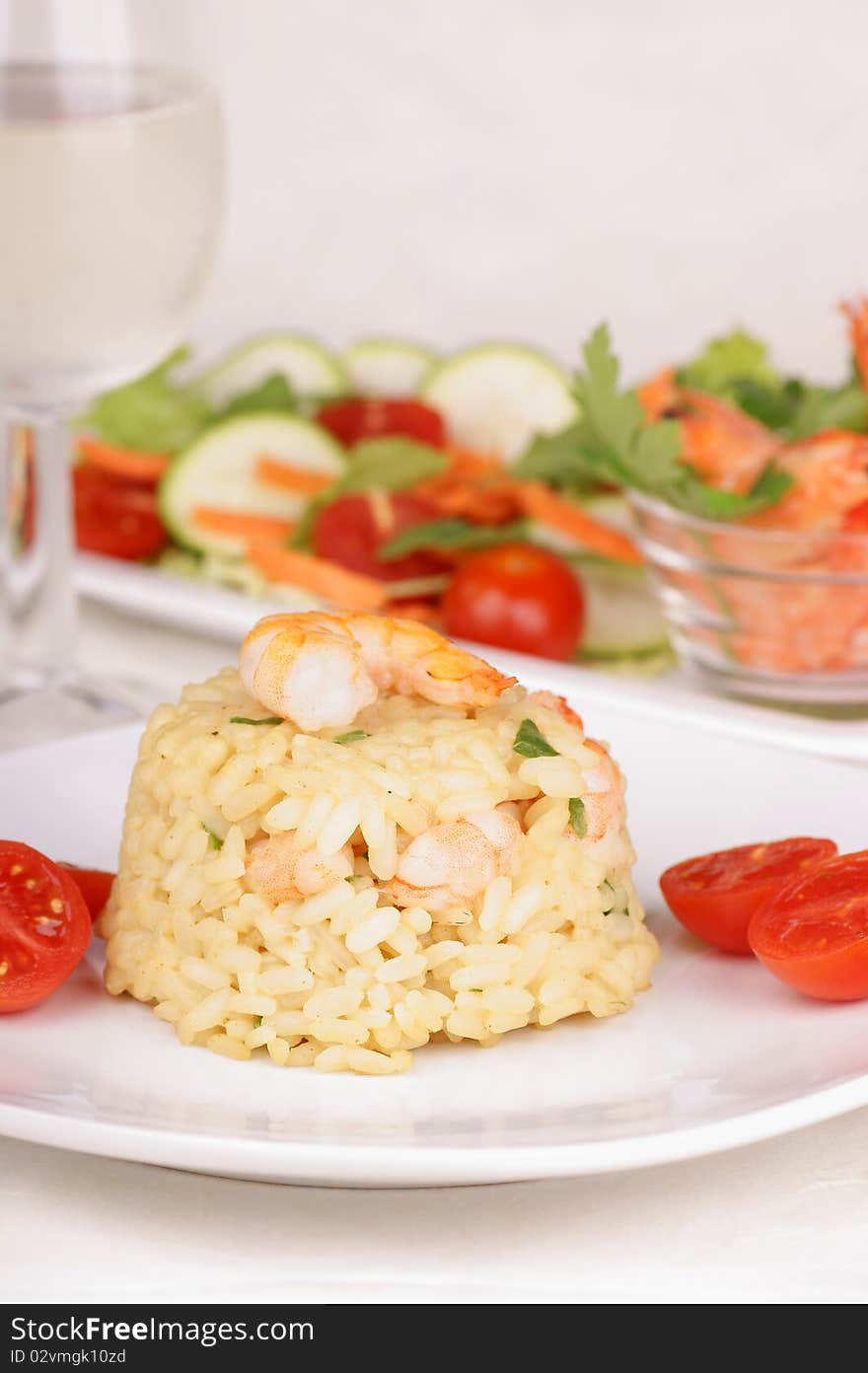 Risotto with shrimps served on a white plate and decorated with cherry tomatoes. Shrimps, mixed salad and a glass of white wine in the background. Selective focus, extremely shallow DOF.