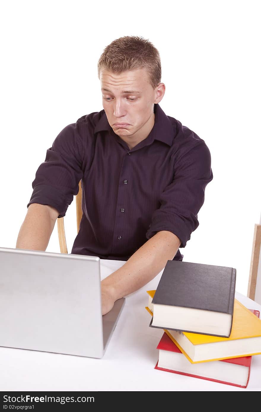 A young man sitting and working on his laptop with a stack of books with a sad expression on his face. A young man sitting and working on his laptop with a stack of books with a sad expression on his face.