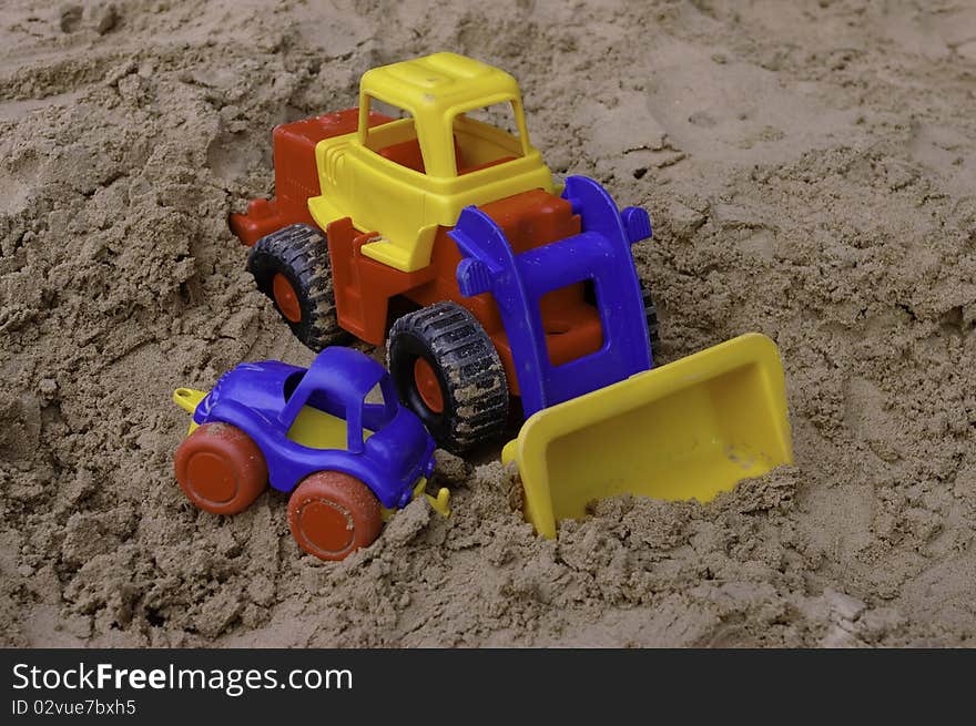 The plastic toy tractor with the small blue car on the sandbox. The plastic toy tractor with the small blue car on the sandbox